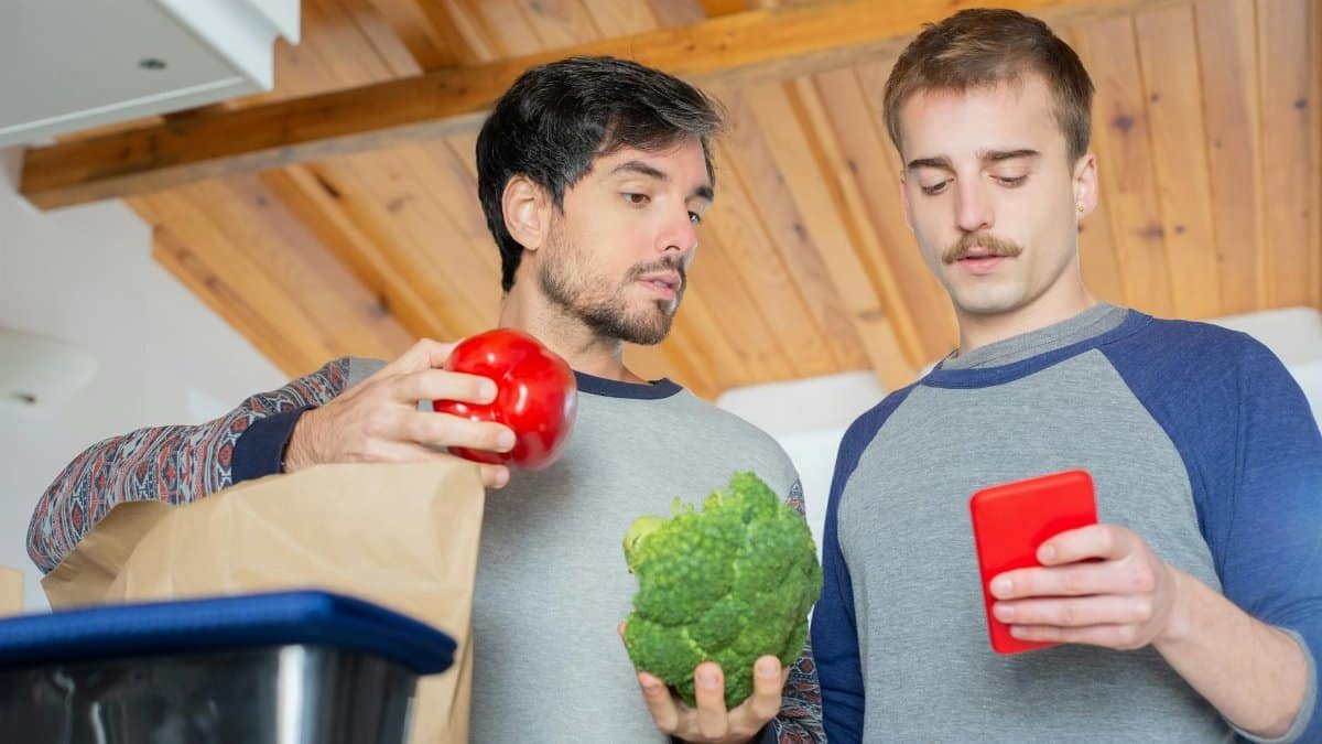 Two men organizing groceries in a kitchen while using a smartphone.
