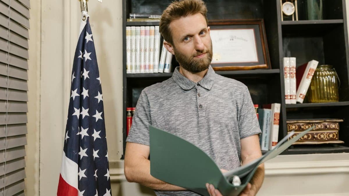 Business professional standing confidently in office holding documents near American flag.