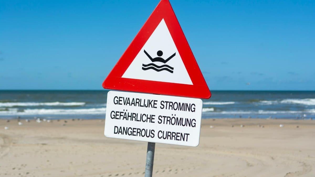 Triangular warning sign on a beach indicating dangerous ocean currents in multiple languages.