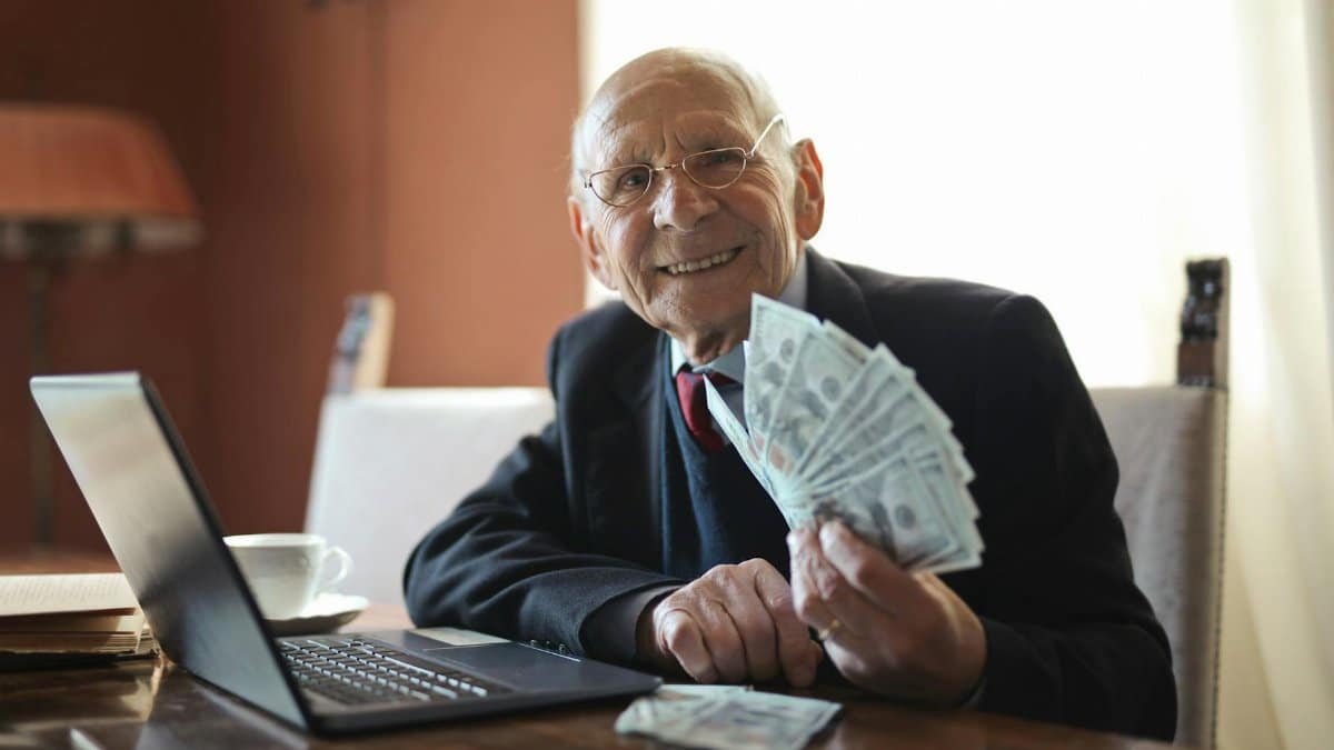 Positive senior businessman in formal suit and eyeglasses holding bunch of bills while working on laptop at table and looking at camera