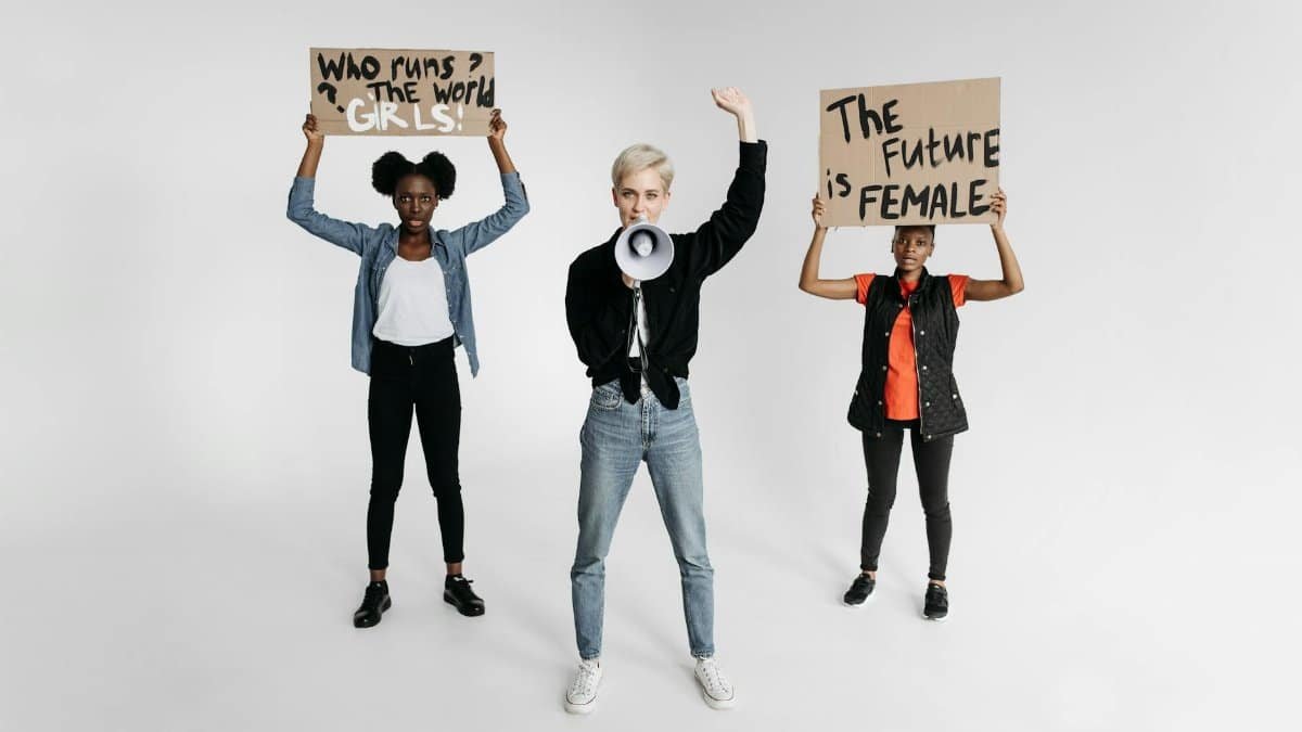 Three women holding feminist protest signs with powerful messages on a white background.