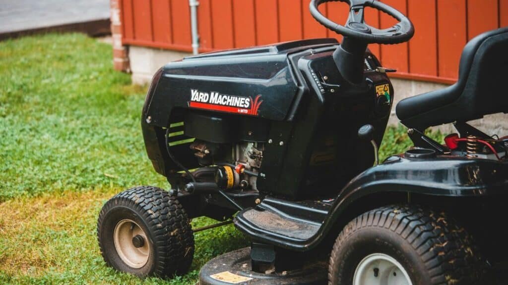 Close-up of a black riding lawn mower parked on a lawn next to a red building.