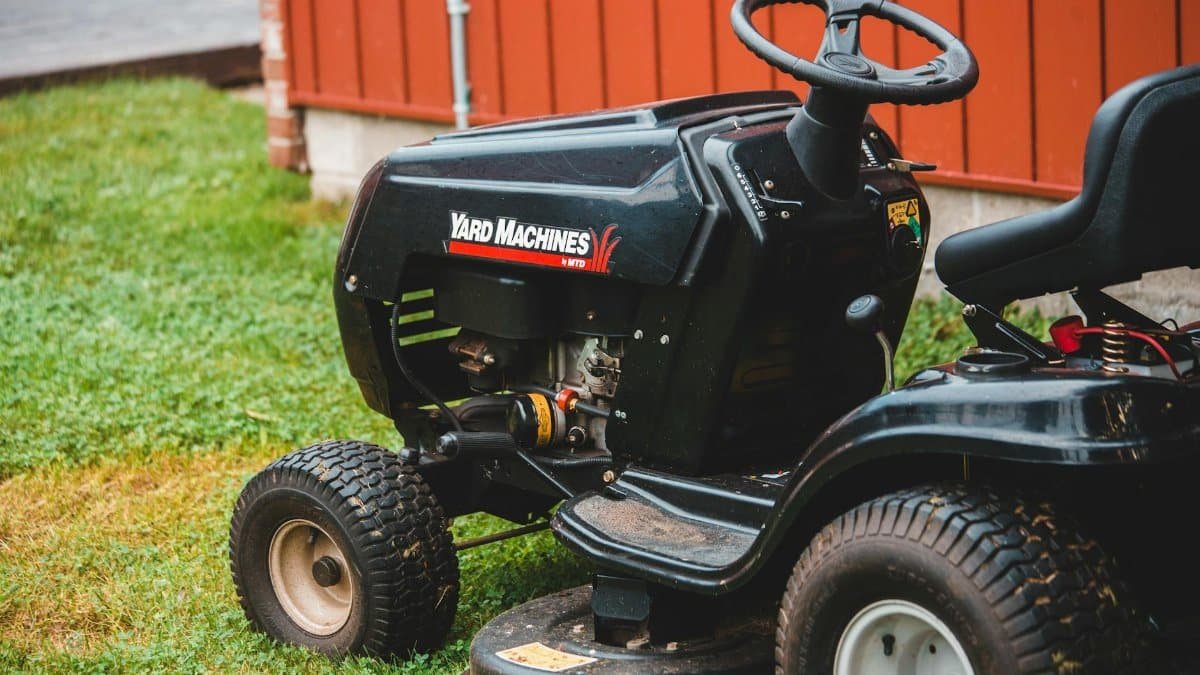 Close-up of a black riding lawn mower parked on a lawn next to a red building.