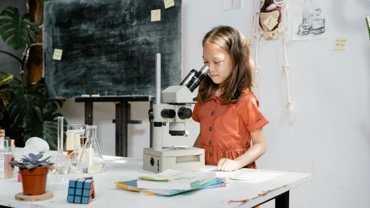 Curious girl observes through microscope in a lively classroom setting, fostering early science education.