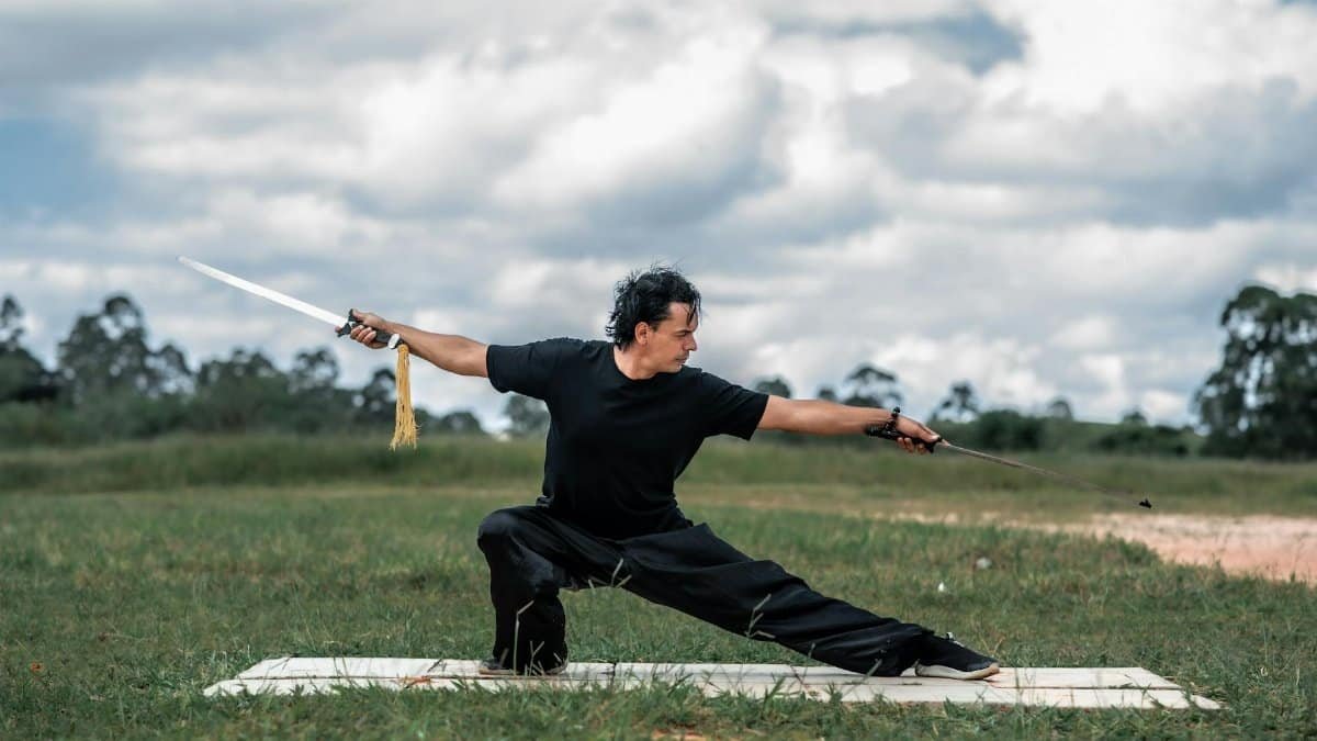 Adult martial artist in black training with swords on grassy field.