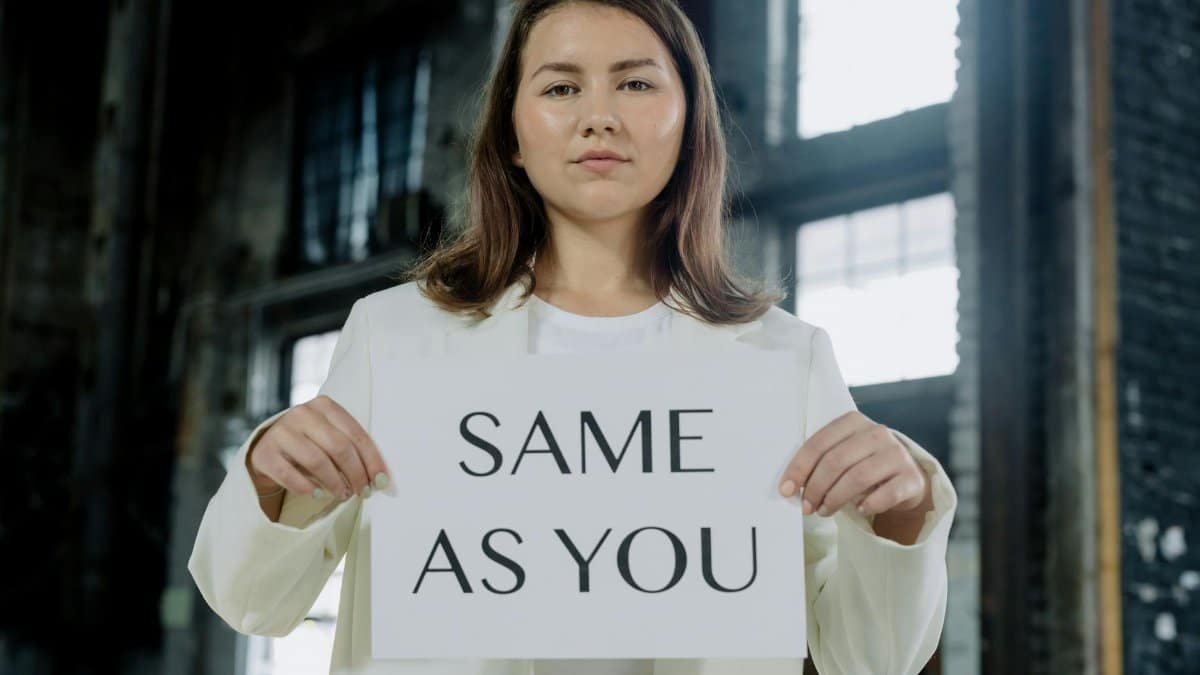A woman in a white outfit holds a 'Same as You' sign indoors, emphasizing equality.
