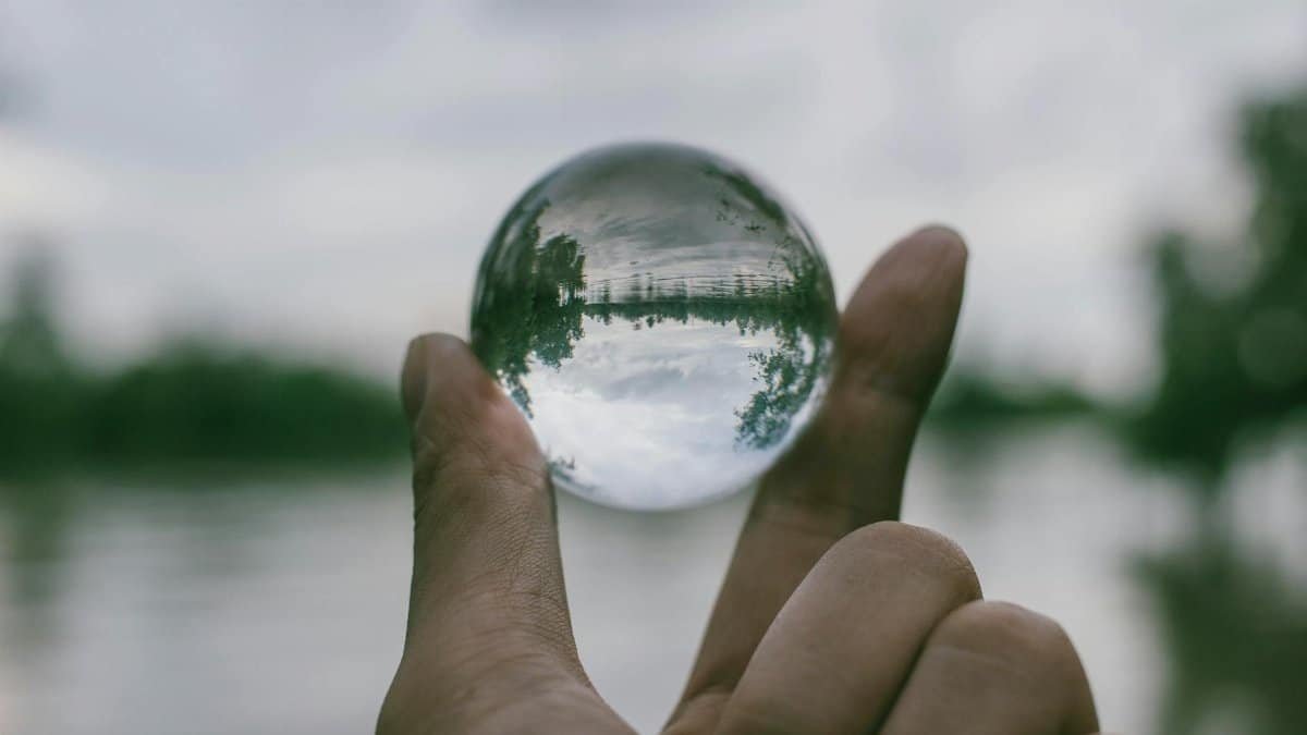 A hand holding a transparent glass sphere outdoors, showcasing an inverted reflection of the landscape.