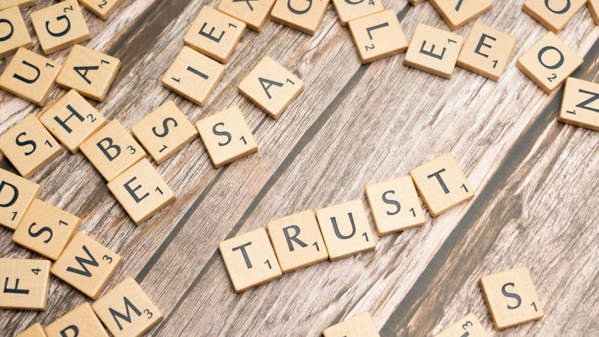 Scrabble letter tiles form the word 'Trust' on a wooden table, surrounded by scattered tiles.