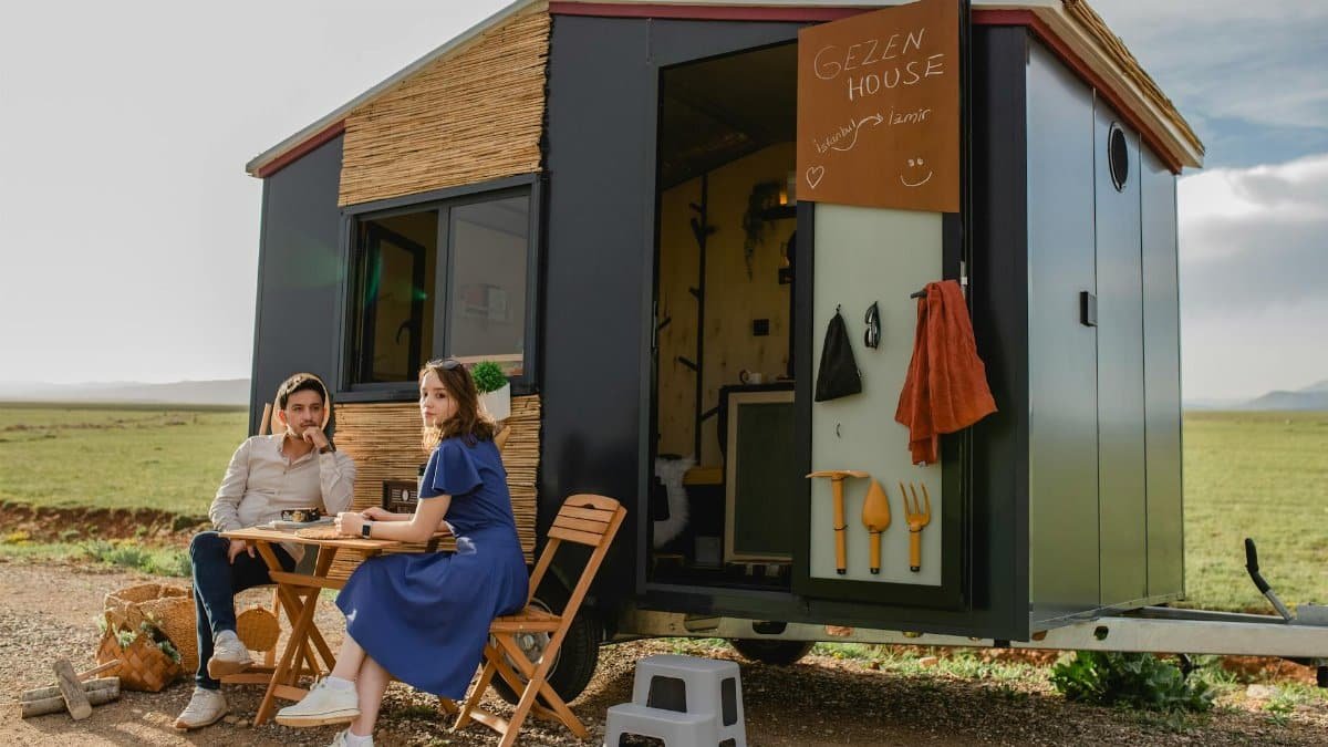 A couple enjoys a relaxing day outdoors near a modern tiny house with chairs and a table.