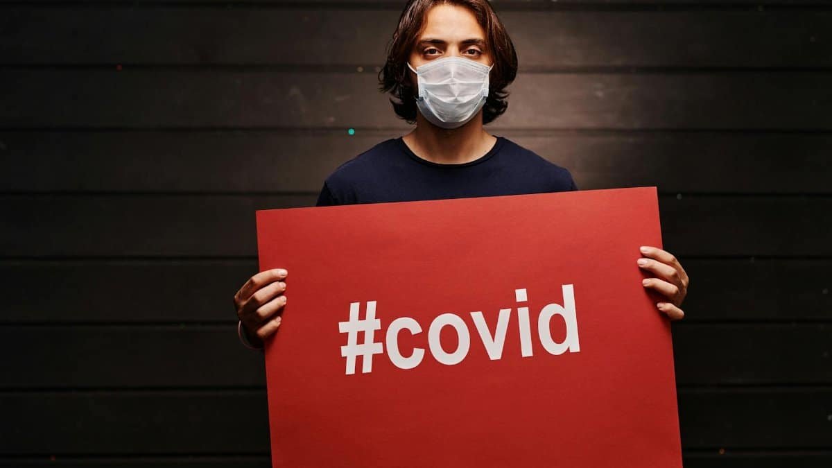 A man in a face mask holds a red sign with '#covid' indoors. Pandemic awareness.
