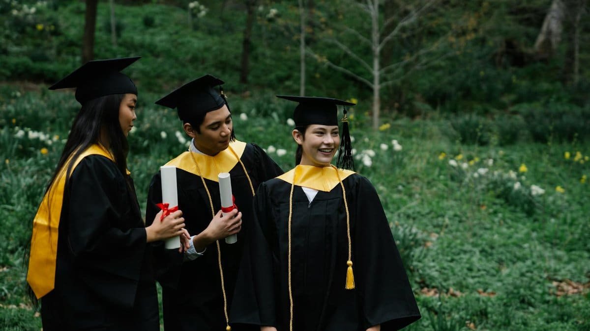 Three graduates in gowns and caps celebrating outdoors among greenery.