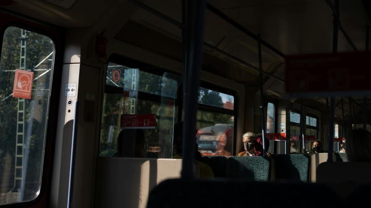Commuters wearing face masks on a sunny urban tram during daytime.