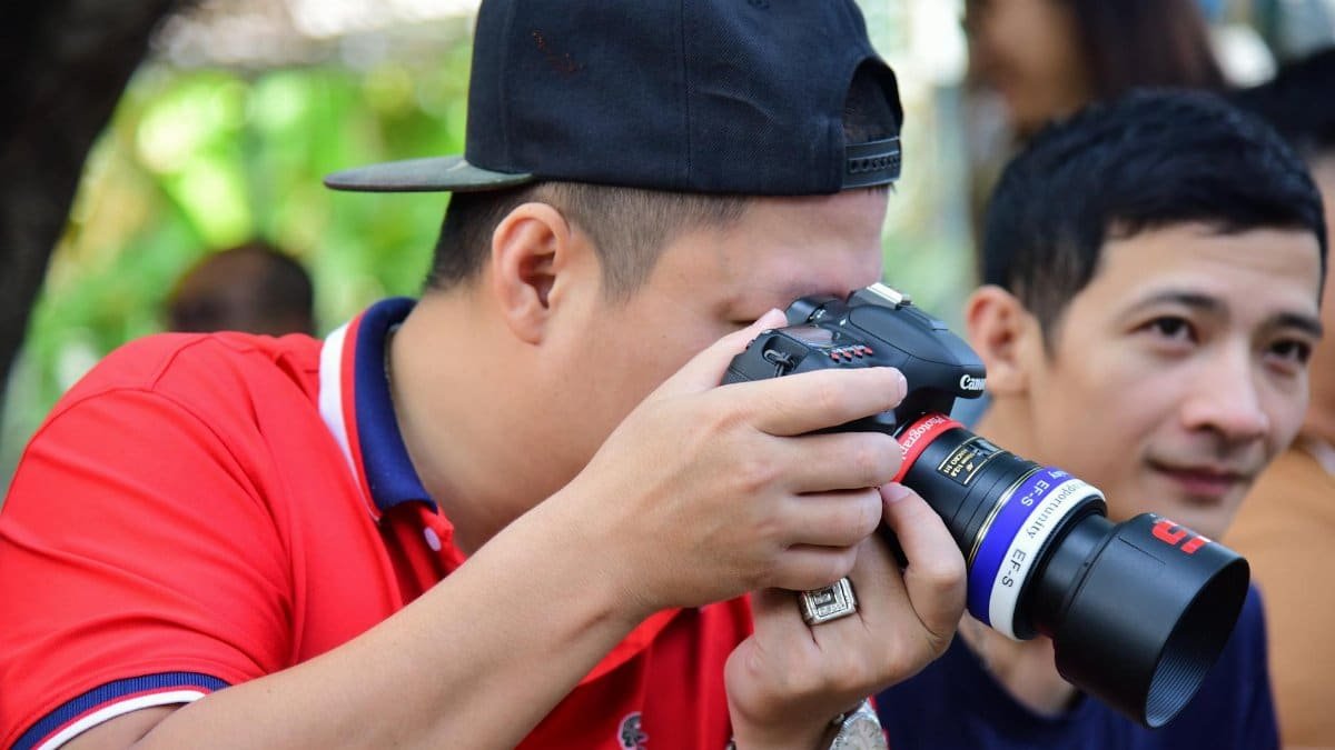 A photographer in a red shirt focuses intently while capturing a moment using a DSLR camera outdoors.