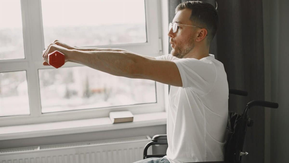 A man in a wheelchair exercising with a dumbbell by a bright window, promoting inclusivity and health.