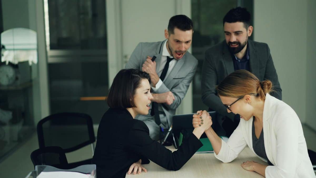 Two female colleagues arm wrestling in an office setting with male coworkers cheering.