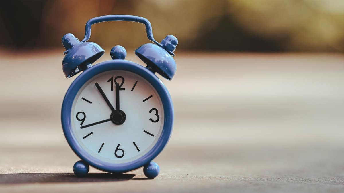 A classic blue alarm clock placed outdoors, displaying five minutes to twelve.