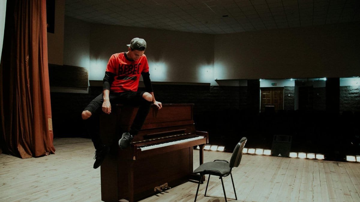 A man in casual attire sits on a piano in an empty concert hall, conveying a sense of rebellion.