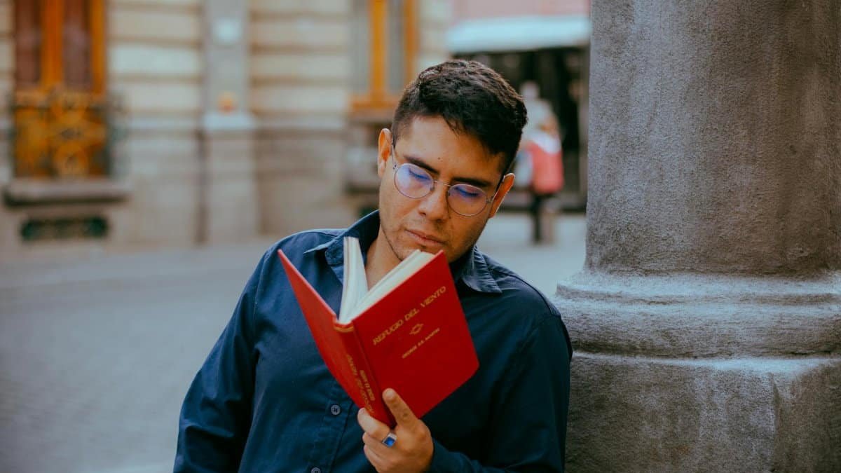 A man in glasses reading a red book while leaning against a column in a city street.