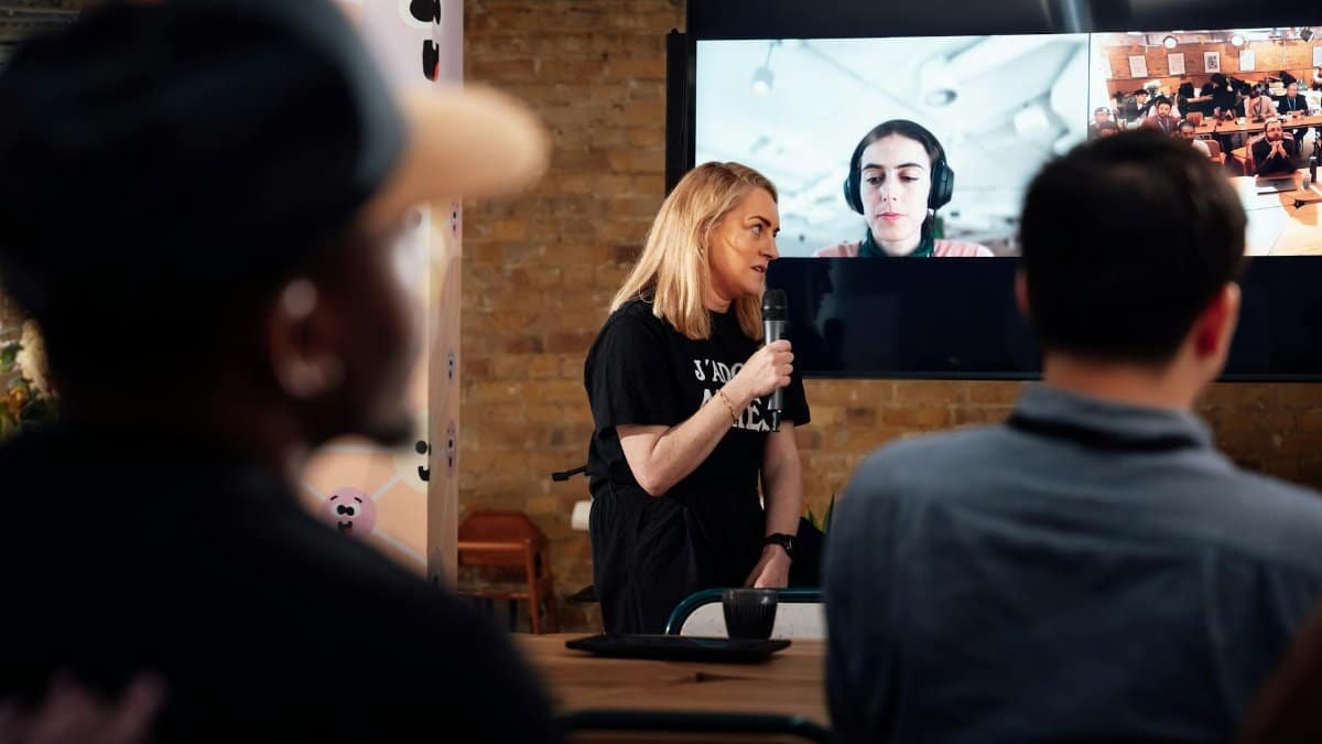 A woman presents to an audience during a hybrid conference in London, with virtual participants on screen.