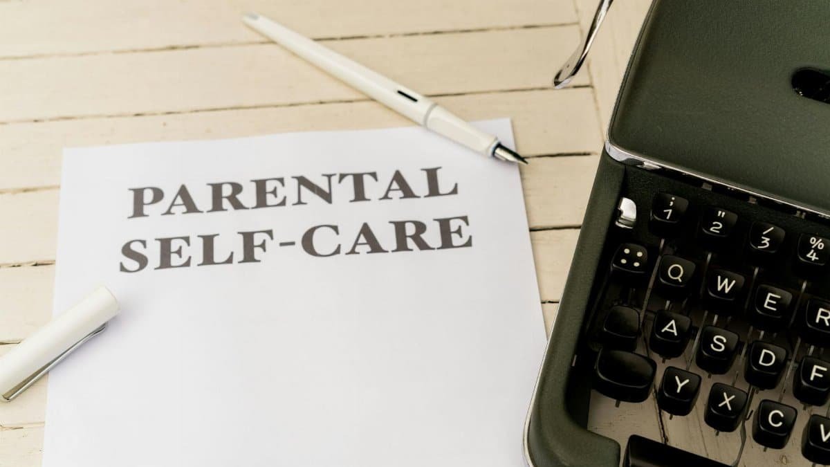 Close-up of paper with 'Parental Self-Care' beside a typewriter, pen, and clip on a wooden desk.