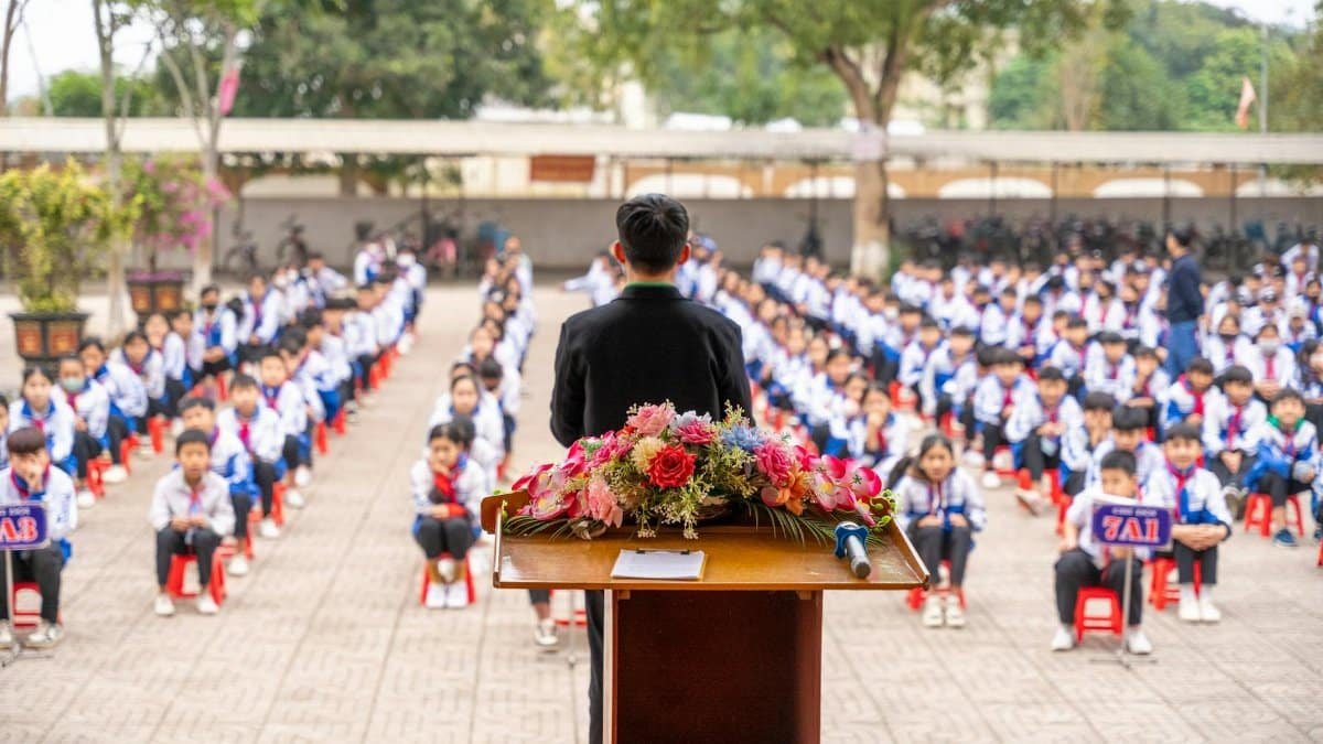 A teacher addresses students seated in an outdoor assembly at a school.