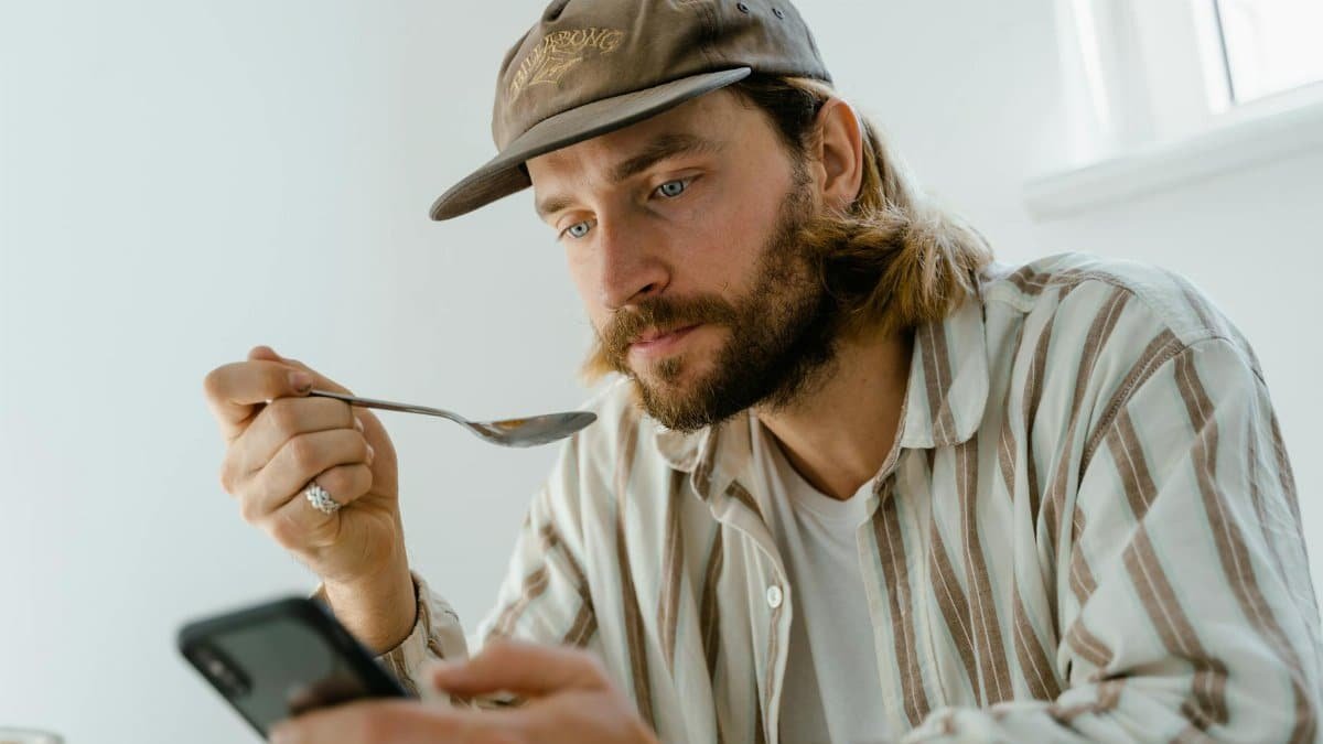 Bearded man wearing cap uses smartphone while eating at home.