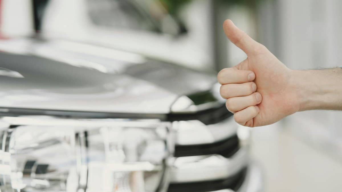 Close-up of a thumbs up gesture next to a car headlight, symbolizing approval or satisfaction.