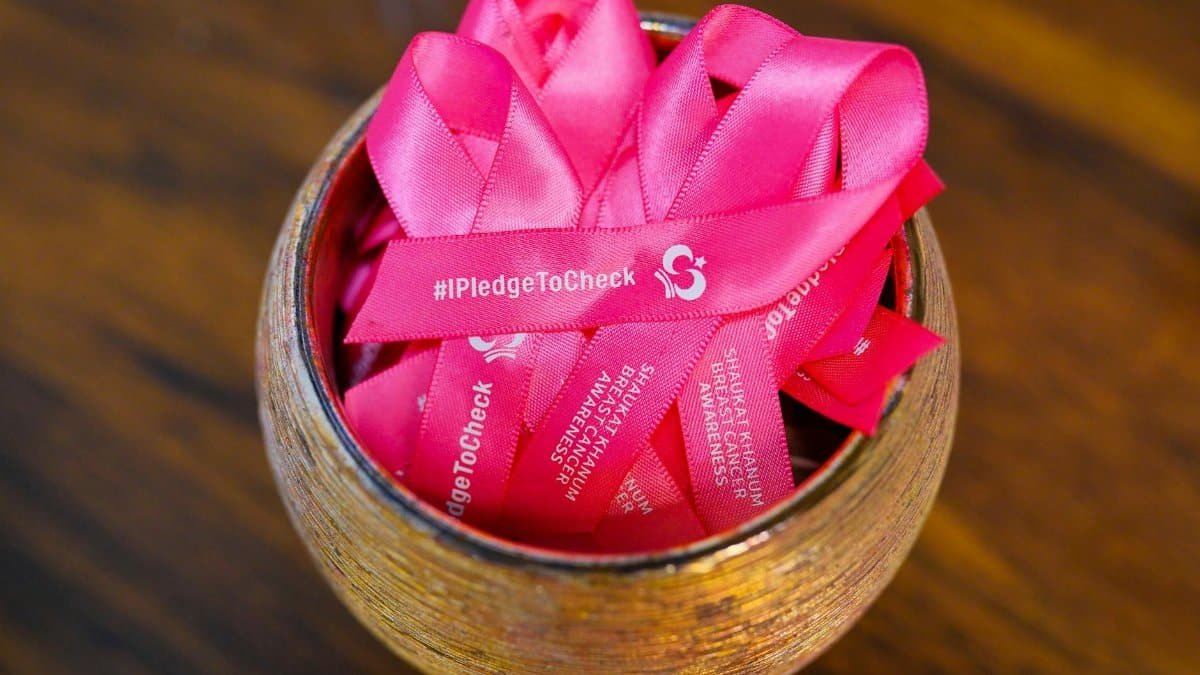 Pink ribbons for breast cancer awareness in a wooden bowl, promoting #IPledgeToCheck initiative.