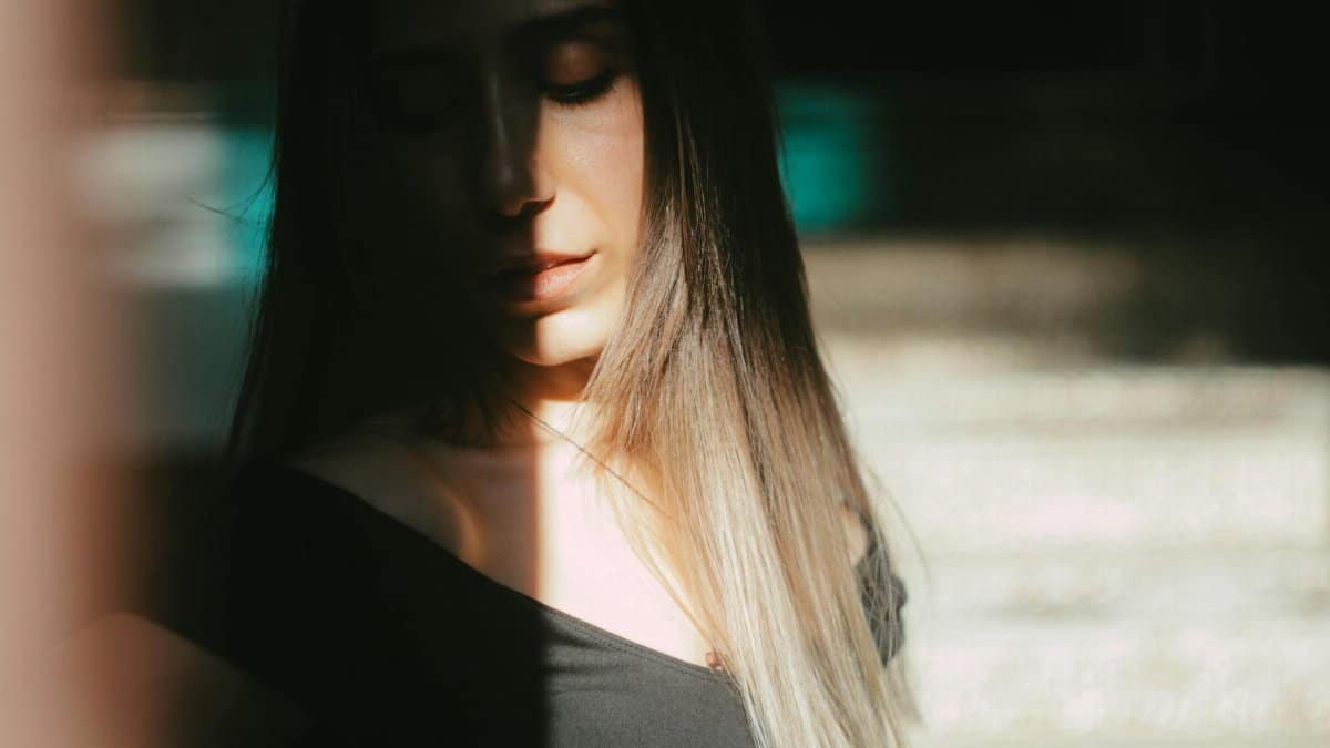 Unrecognizable female with dark hair standing with closed eyes under sunlight covering face while standing on street with blurred background