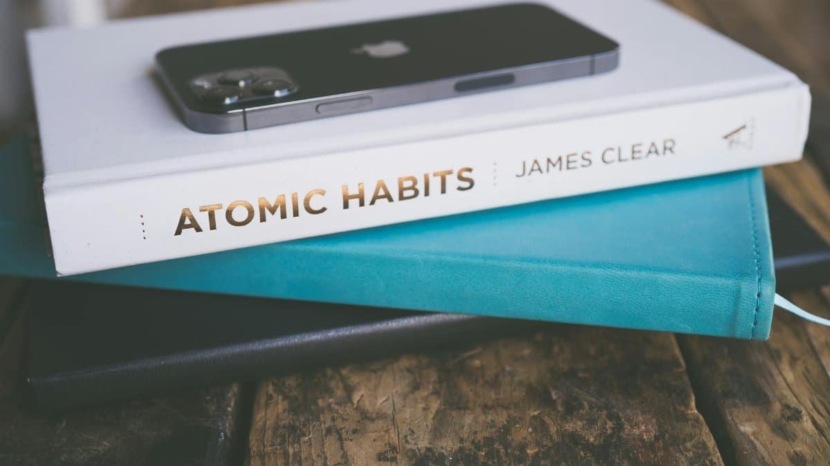 Close-up of a smartphone on a book stack, featuring 'Atomic Habits' by James Clear on a wooden surface.