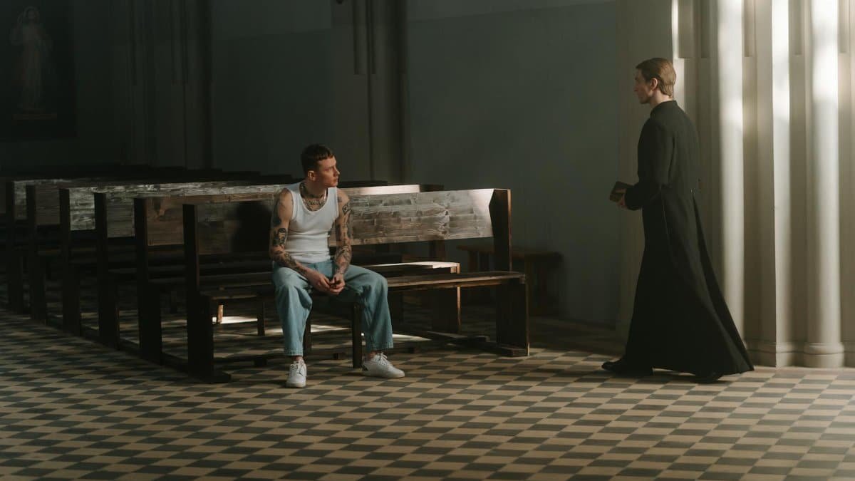 A tattooed man and a priest in a spiritual encounter within a church setting.