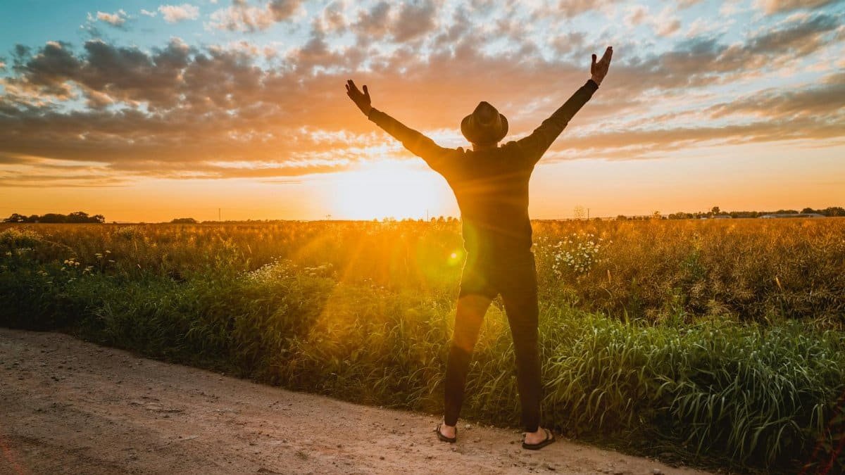 A man raises arms in freedom at sunrise in a rural field, expressing joy and connection with nature.