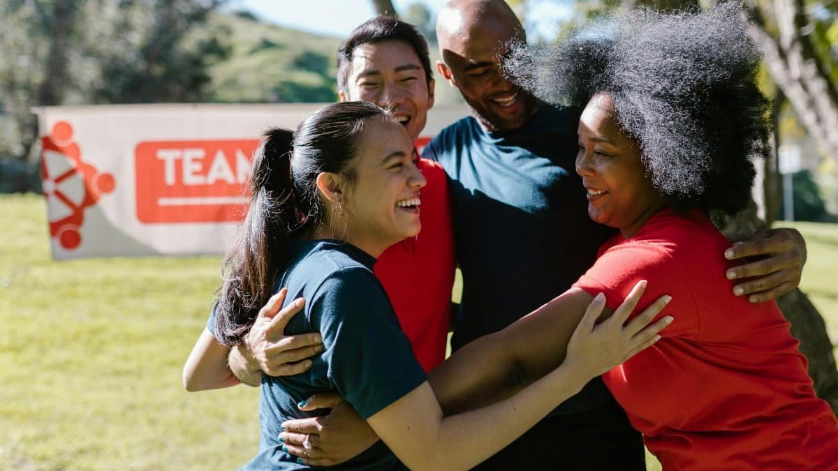 A joyful, diverse group embraces during an outdoor team building event in a sunny park.