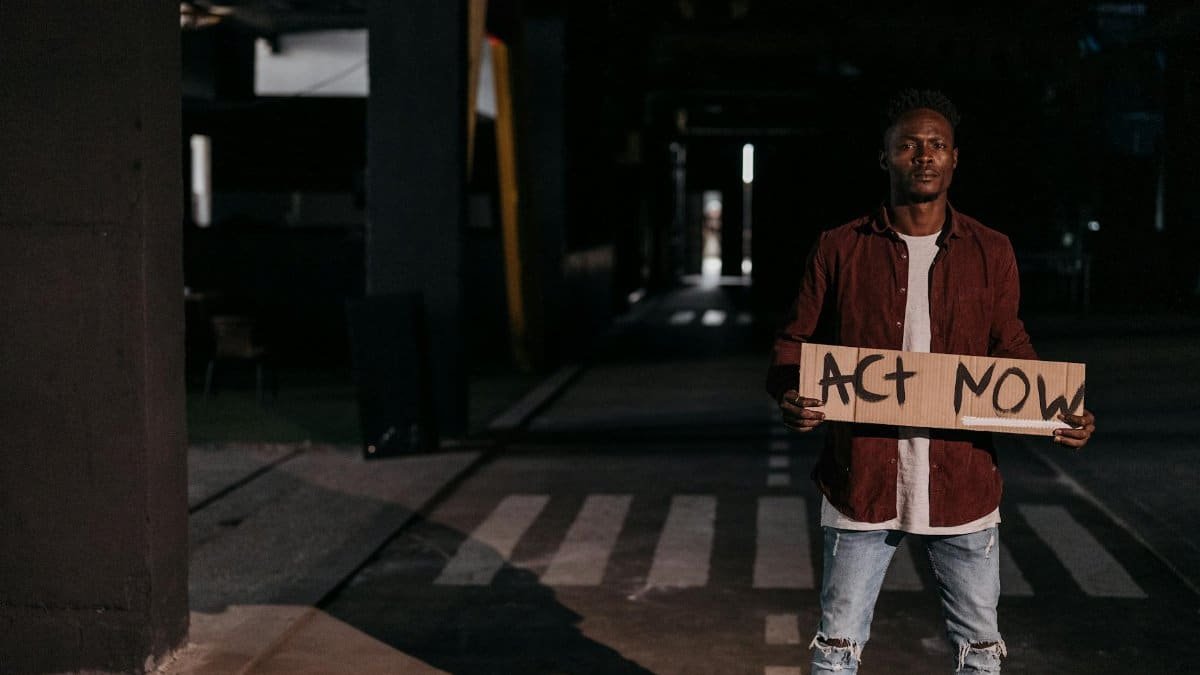 A man stands on a street at night holding a cardboard sign reading 'Act Now,' highlighting a call to action.