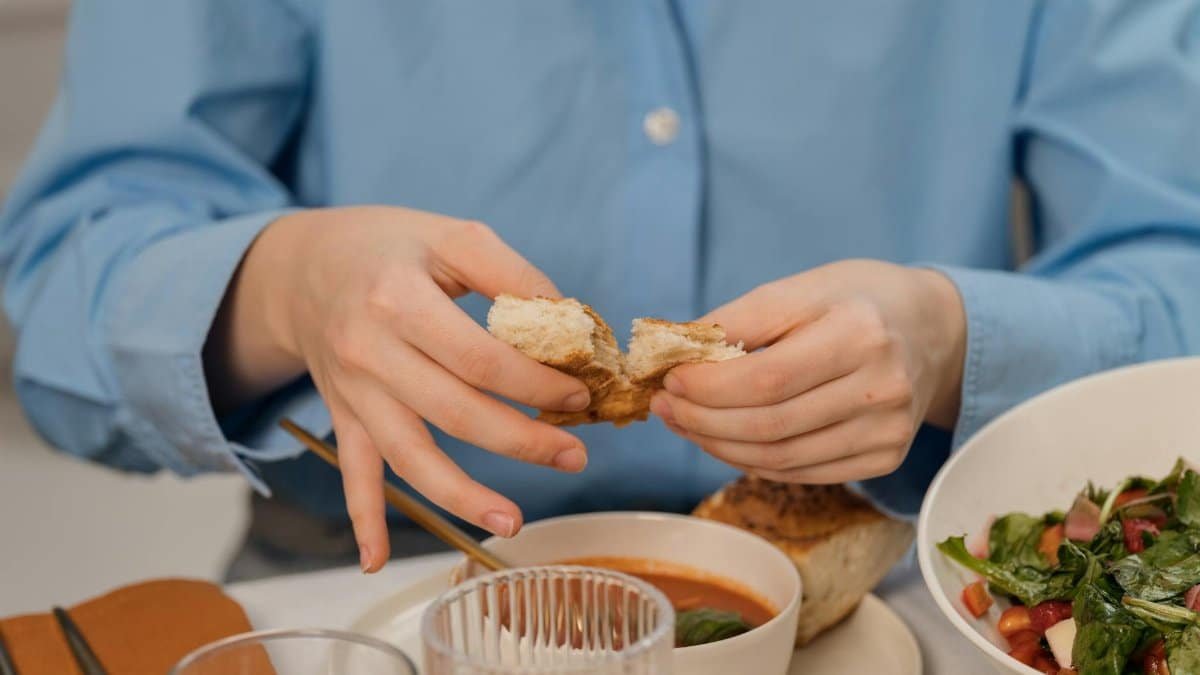 A woman tearing bread over a bowl of soup, wearing a blue shirt, indoors.