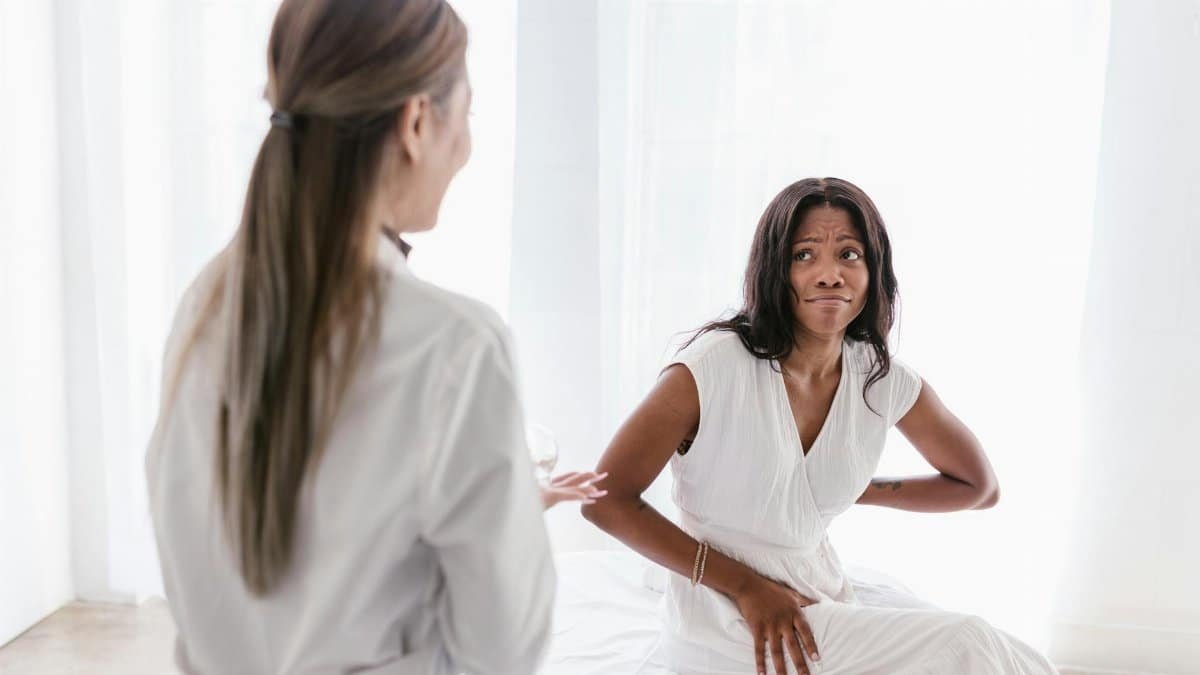 A woman with back pain consults her doctor in a clinical setting wearing a white dress.