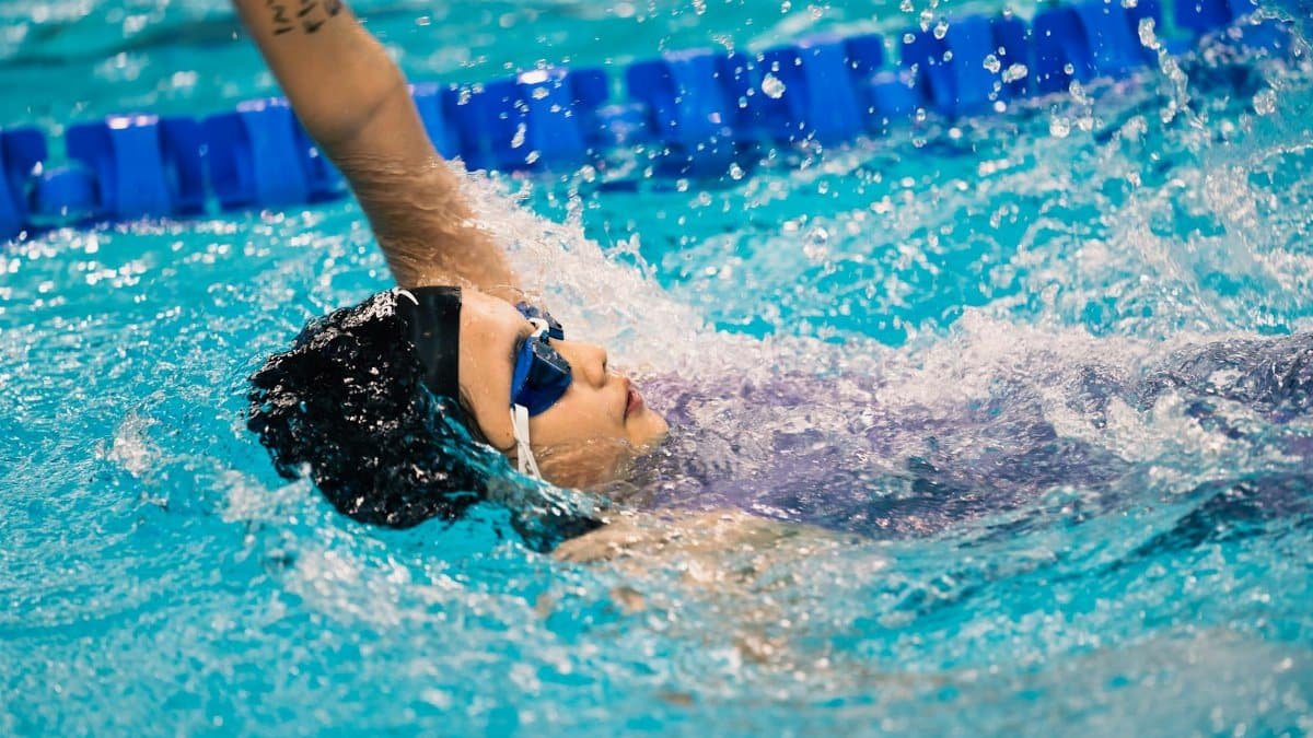 Swimmer doing backstroke in pool with blue goggles during a competitive event.