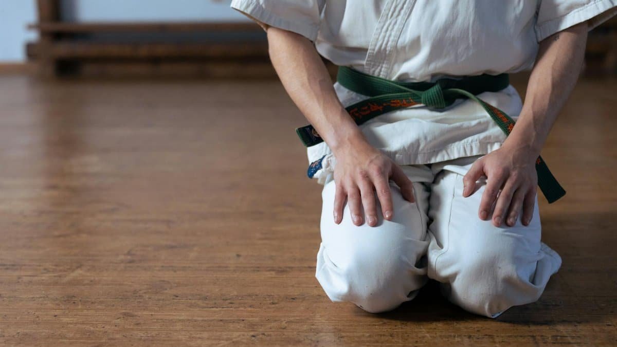 A martial artist in a white uniform kneels on a wooden dojo floor, focusing on practice.