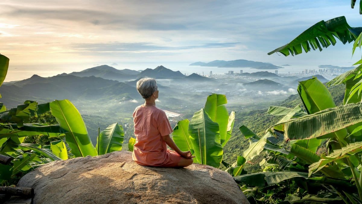 Elderly person meditating on a rock with panoramic tropical mountain view and banana trees.
