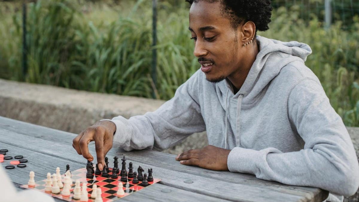 African American chess player playing board game while sitting at table in park with friend