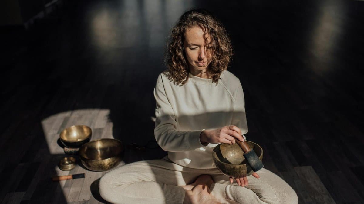 Female sound therapist sitting with crossed legs on parquet while playing singing bowl with mallet during meditation practice
