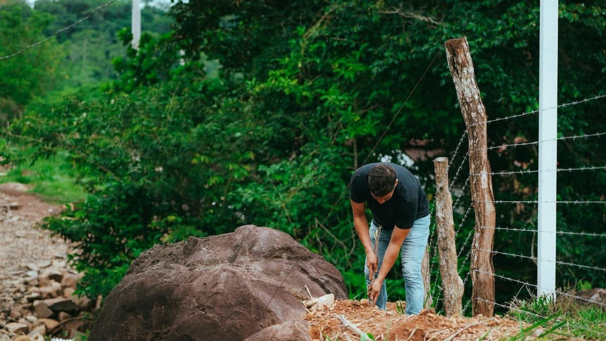 A man working near a barbed wire fence in a natural landscape, embodying outdoor labor.