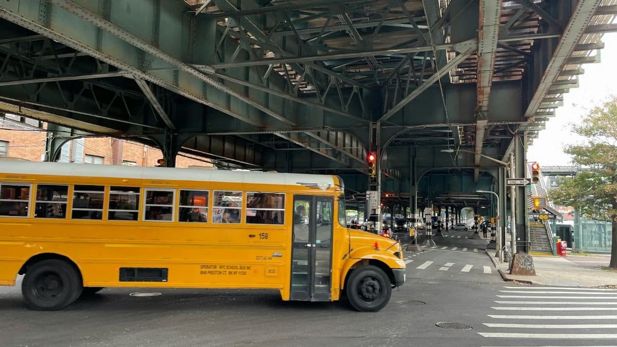 A yellow school bus drives under a New York overpass on a daytime street.