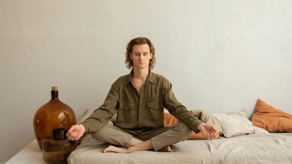 Full length young relaxed male in casual wear sitting with eyes closed in Lotus Pose on soft mattress and meditating