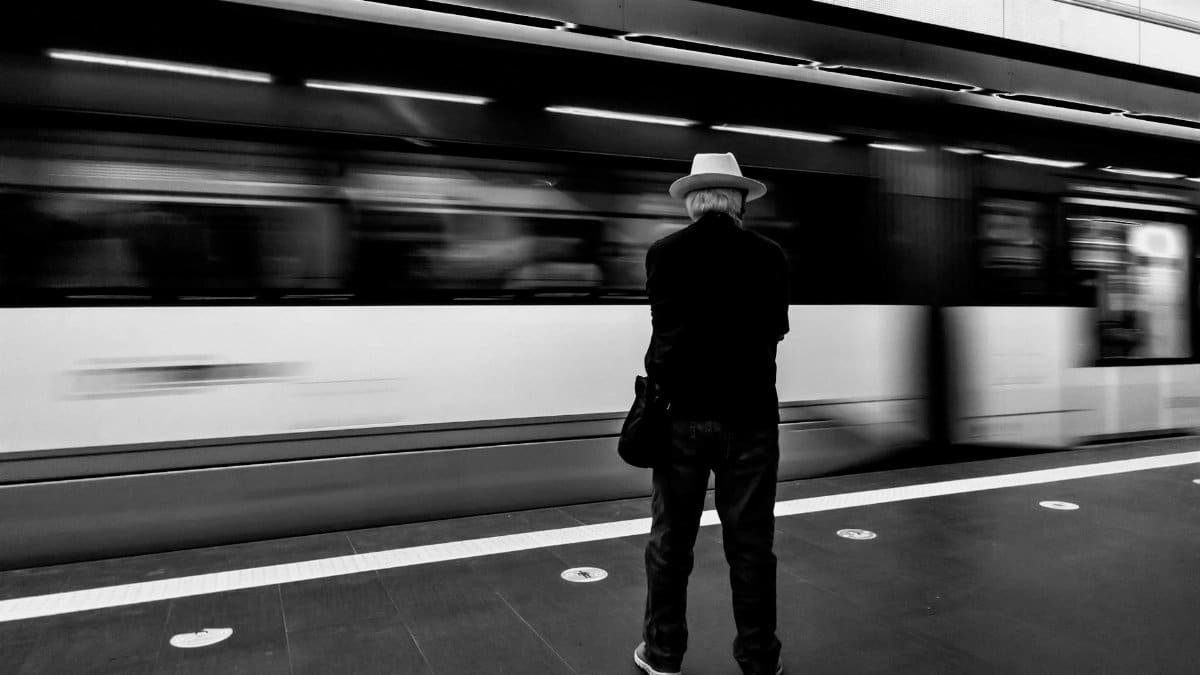 Man in hat waiting at Alicante train station, blurred train passing by, monochrome.