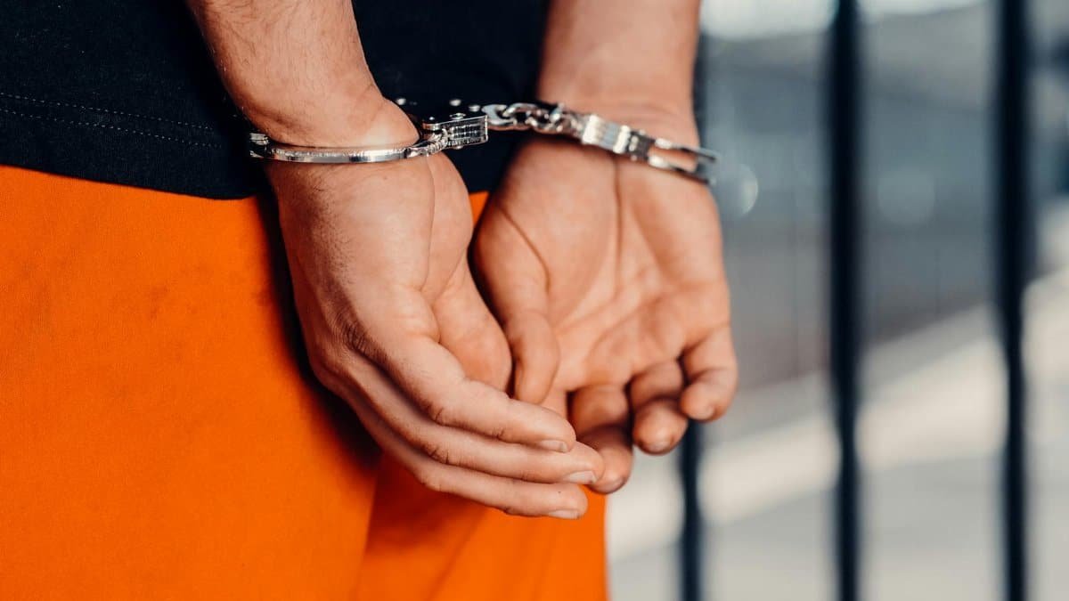 Close-up of person in handcuffs wearing orange attire behind prison bars, symbolizing arrest.