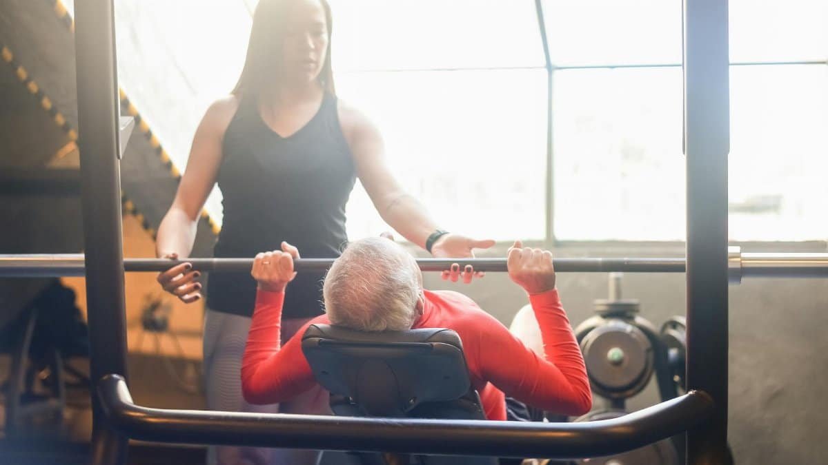 Elderly man in gym performing bench press with female trainer assistance, promoting fitness and health.