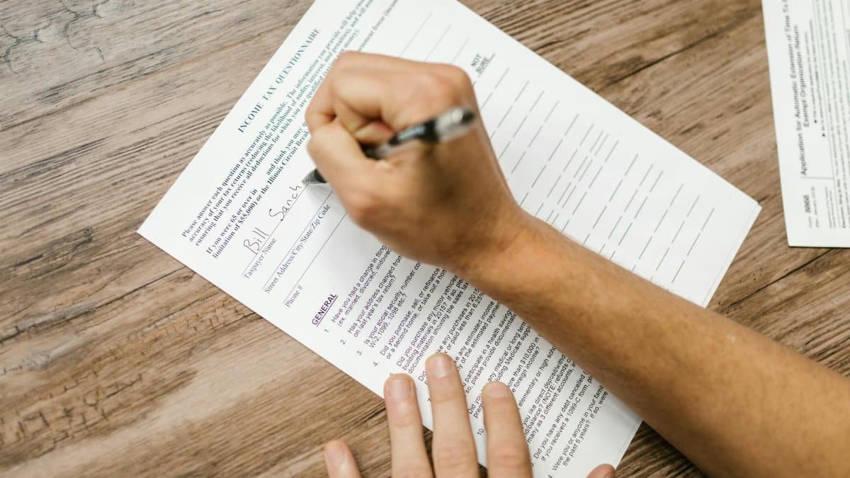 Person filling out a tax questionnaire with a pen, showing close-up on wooden table.
