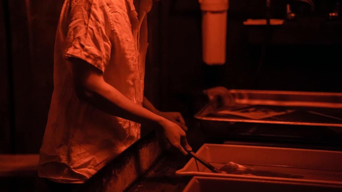 Photographer developing prints in a dimly lit darkroom with red lighting and chemical trays.