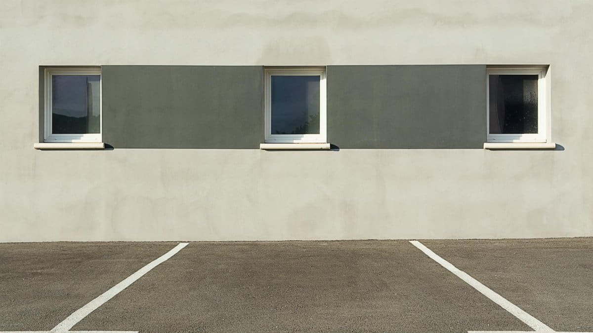 A modern building facade with three windows, featuring a minimalist design and an empty parking lot in the foreground.